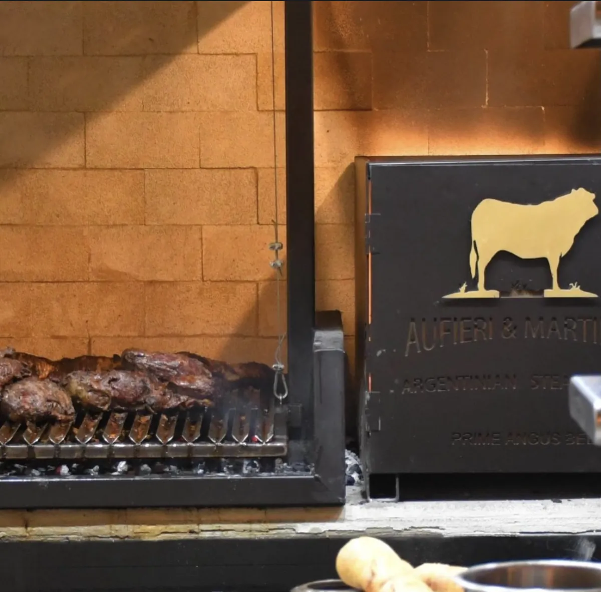 Steaks on the parrilla grill over embers beside the Aufieri & Martínez cow emblem