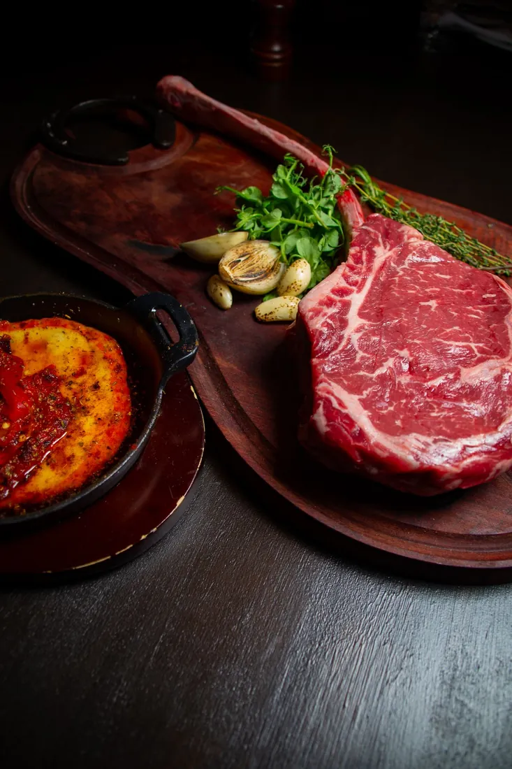 A raw ribeye on a wooden board beside provoleta